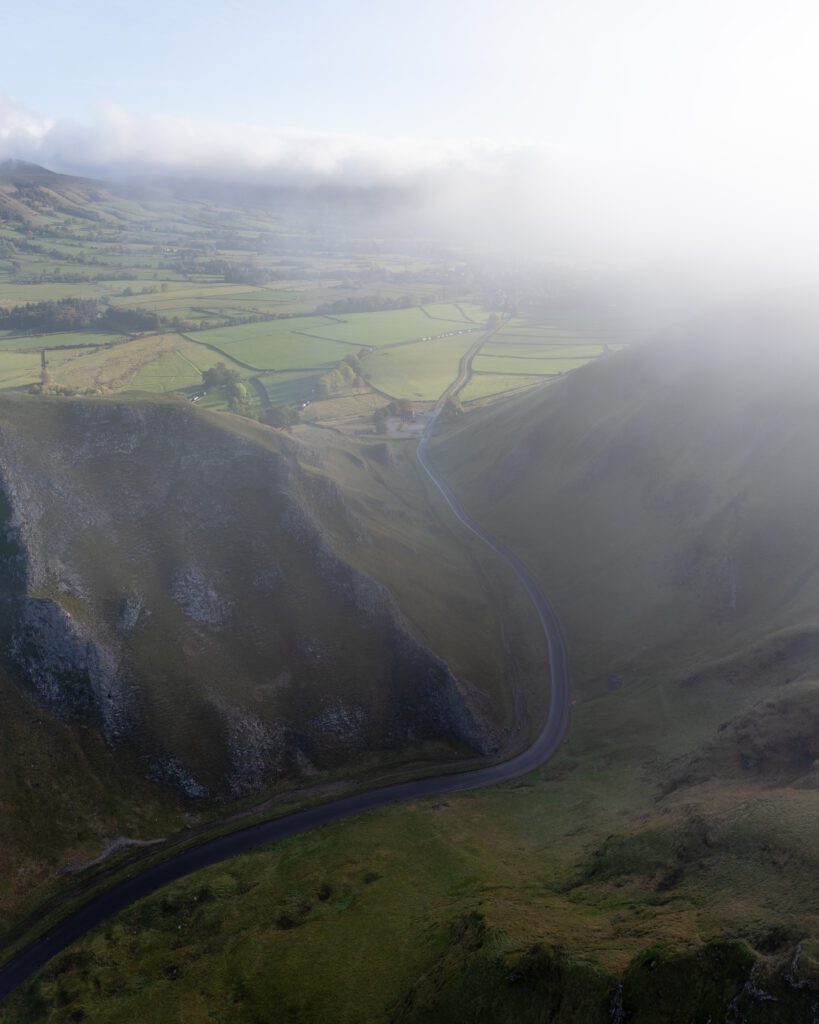 Aerial drone view of Winnats Pass road winding through misty Peak District hills in England.