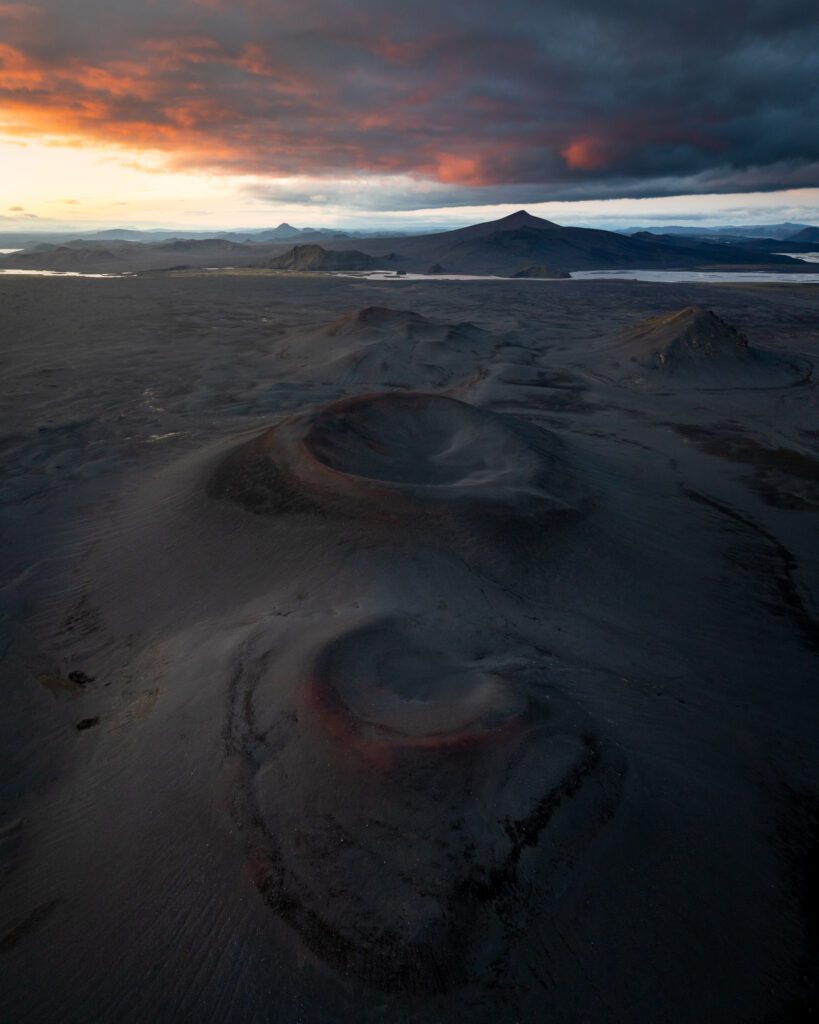 Drone view of twin red-rimmed cinder cones in Fjallabak lit by a fiery sunset on a black ash plain.