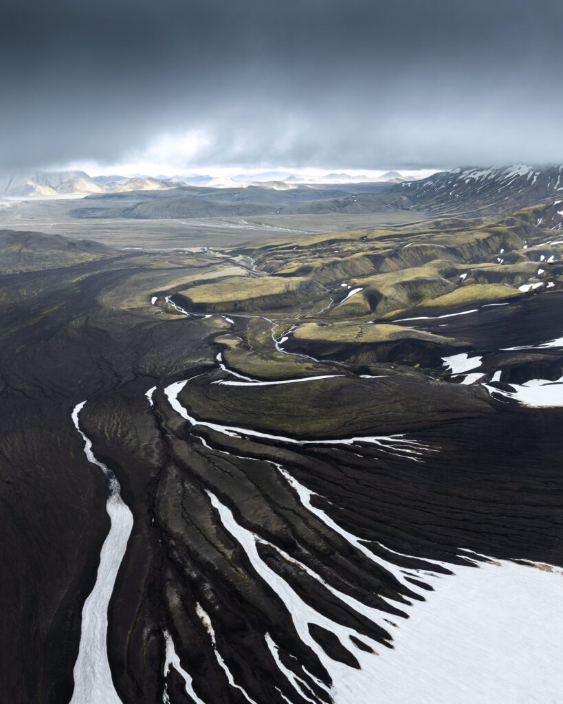 Black streams carving down through a snow-streaked plateau under storm clouds, seen from a drone.