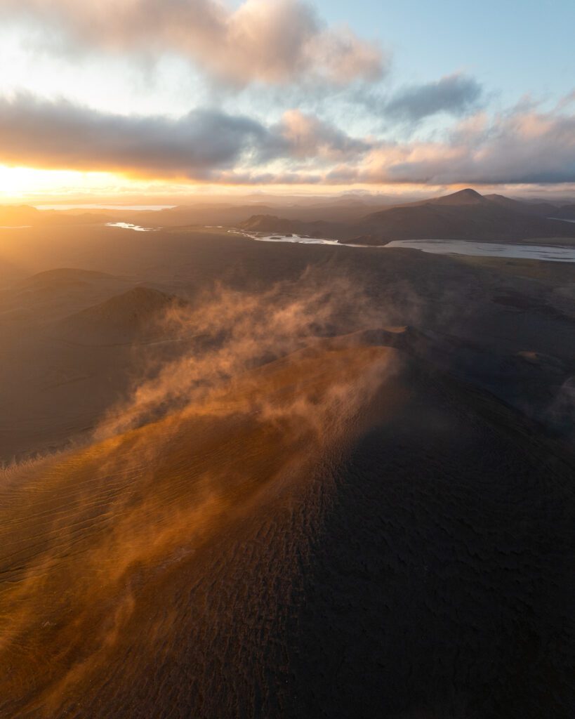 Sunset drone view of golden mist sweeping across a dark volcanic ridge with rivers fading into the distance.