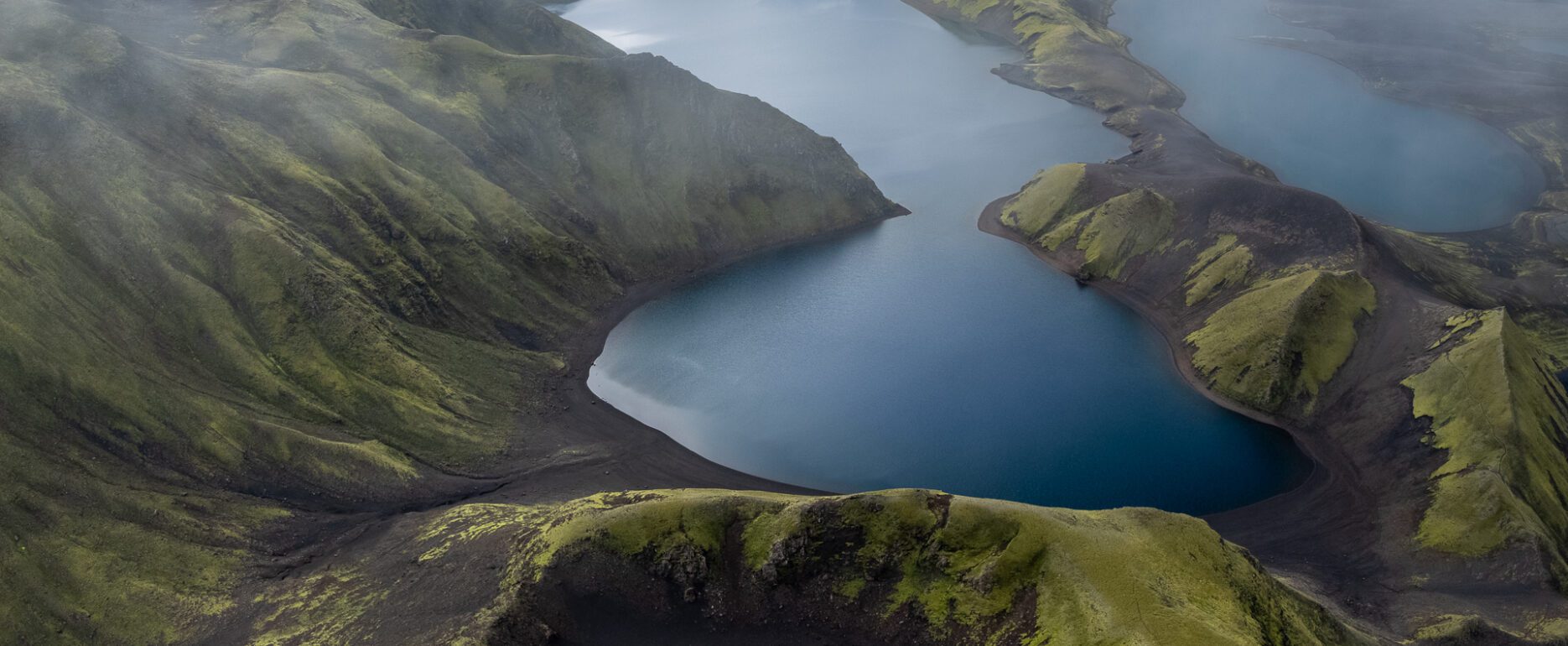 Aerial drone view of a misty lake nestled among green hills and a small crater in the Icelandic Highlands.