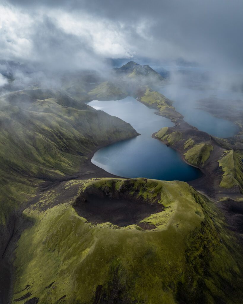 Aerial drone view of a misty lake nestled among green hills and a small crater in the Icelandic Highlands.