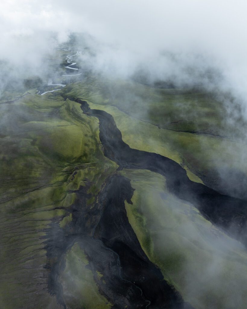Dark streams cutting through green mossy uplands shrouded in heavy mist, viewed from above.