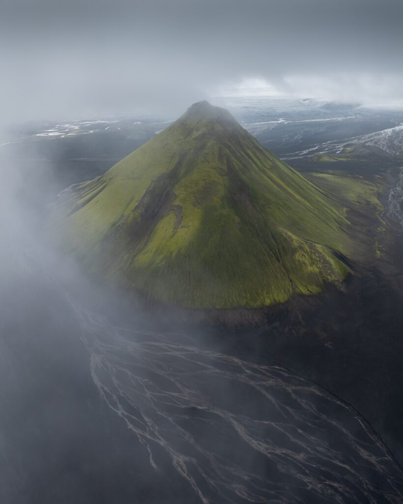 Mælifell rising as a lone moss-clad cone from braided black sand under heavy low cloud.