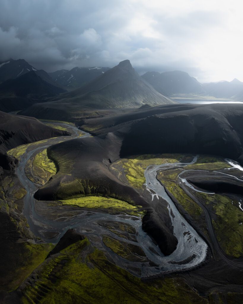 Meandering river winding around mossy mounds under stormy grey skies, viewed from the air.