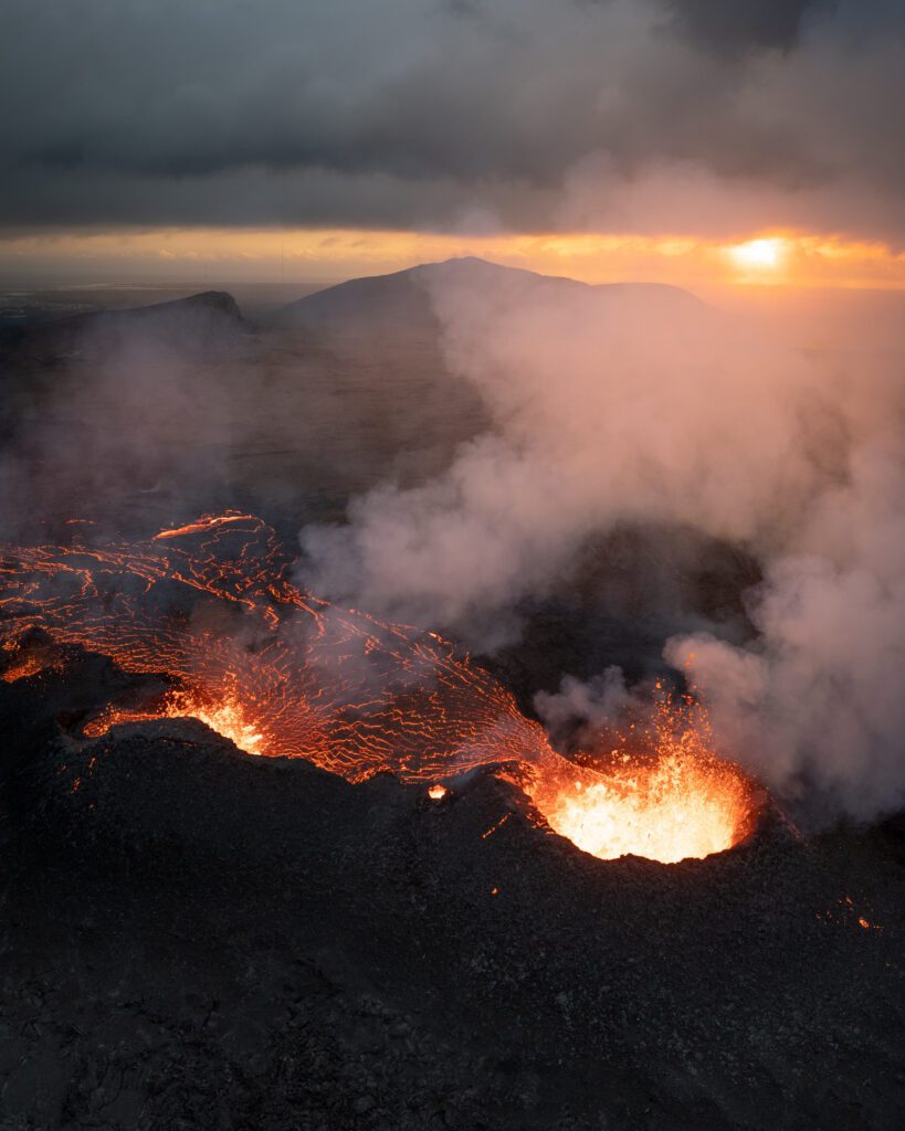 Aerial of an erupting fissure with bubbling lava and steam, distant mountain at sunset.