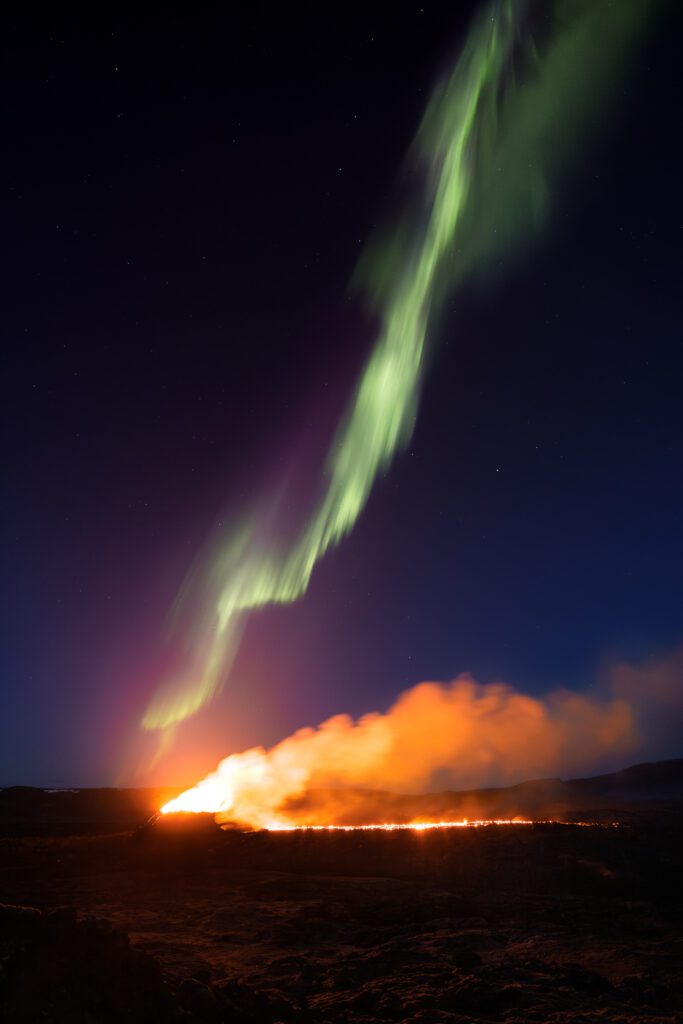 Long fissure eruption glowing at the horizon below a green aurora streaking the night sky.