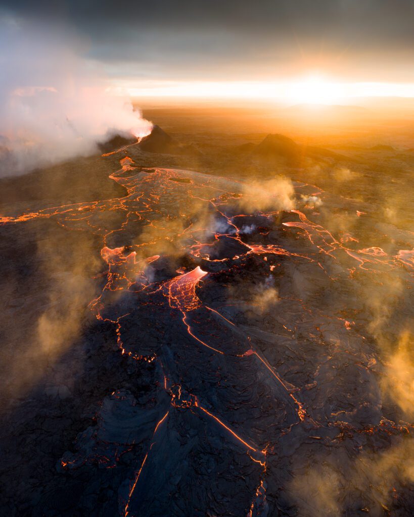 Aerial sunset view of a vast lava field with steam vents stretching to a distant erupting cone.
