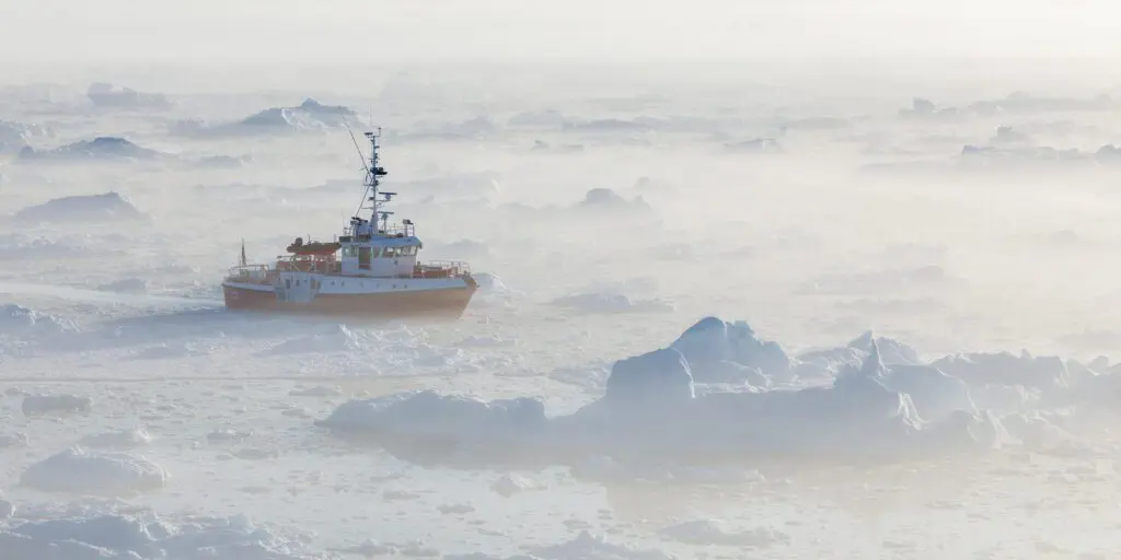 Small expedition boat threading between icebergs in thick sea fog off West Greenland