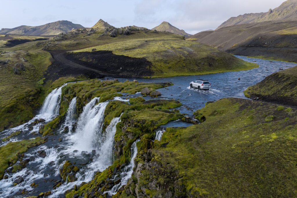 Silver Land Cruiser pushing through a highland river, spray behind it and waterfalls on the mossy bank.