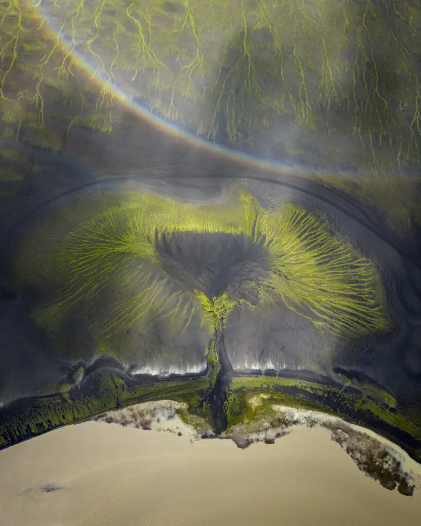 Green moss veins radiating across black sand under a faint rainbow, seen from above